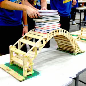 American public High school students have built a bridge out of wooden popsicle sticks The bridge is sitting on a table The students are testing the strength of the bridge by stacking books on top of it The bridge is not built out of books It only ha-1